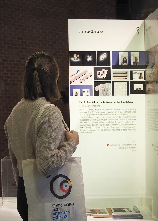 Loida Massanet carrying a "9º Encuentro Bienal Iberoamericana de Diseño - Enseñanza y Diseño" tote bag while observing the "Dentistas Solidarios" project exposition.
